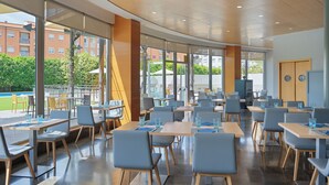Een moderne eetruimte met tafels en stoelen, grote ramen en uitzicht op gebouwen en groen.