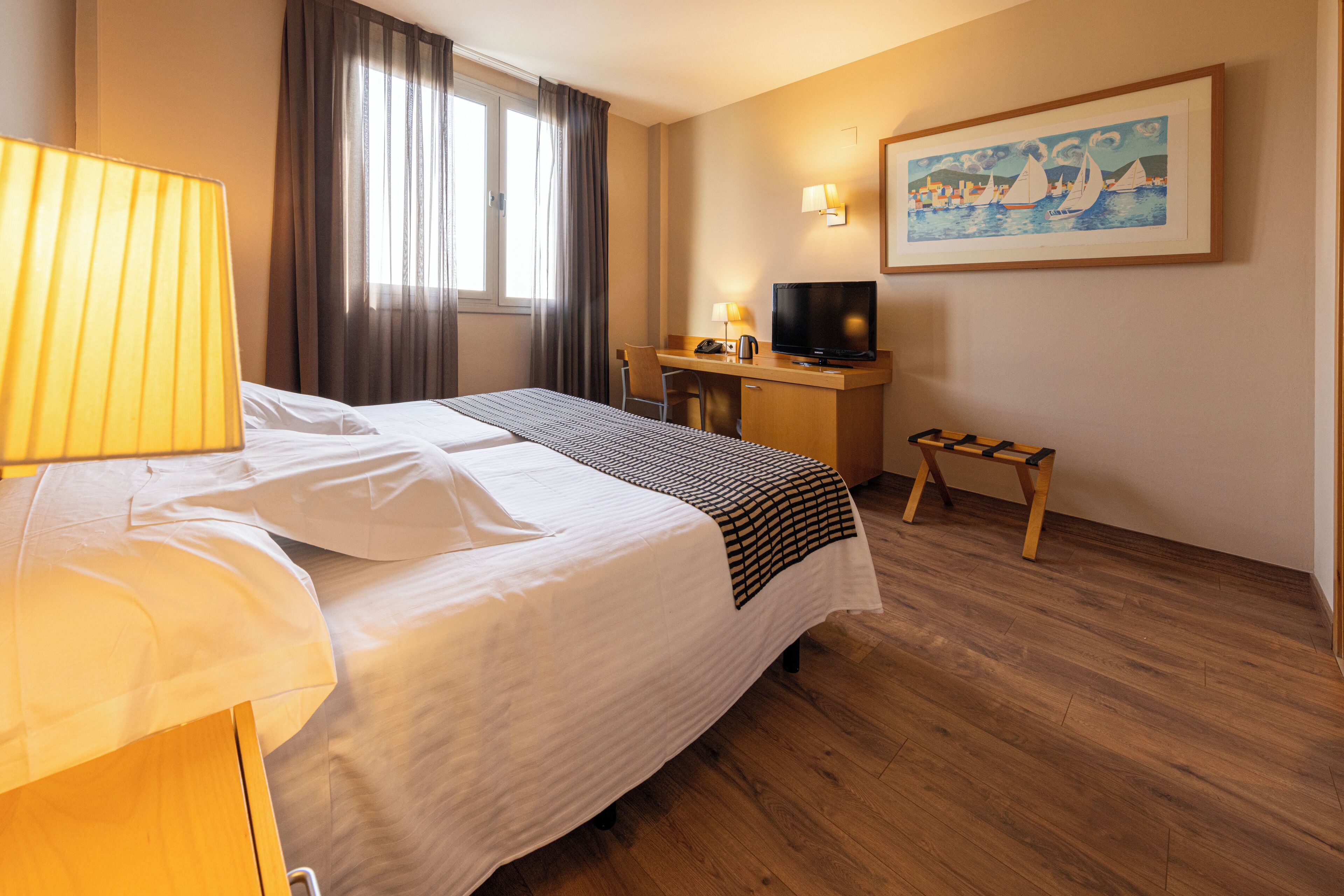 A hotel room with a bed, a desk with a television, a chair, a small table, and a painting on the wall.