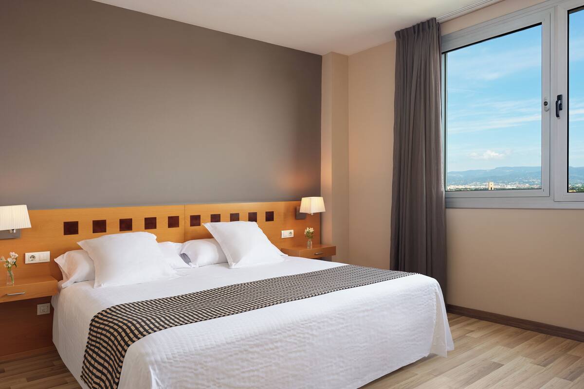 Una cama bien tendida con ropa de cama blanca y un plaid estampado, un cabecero de madera, una mesita de noche con una lámpara y una ventana con vistas a un paisaje urbano.