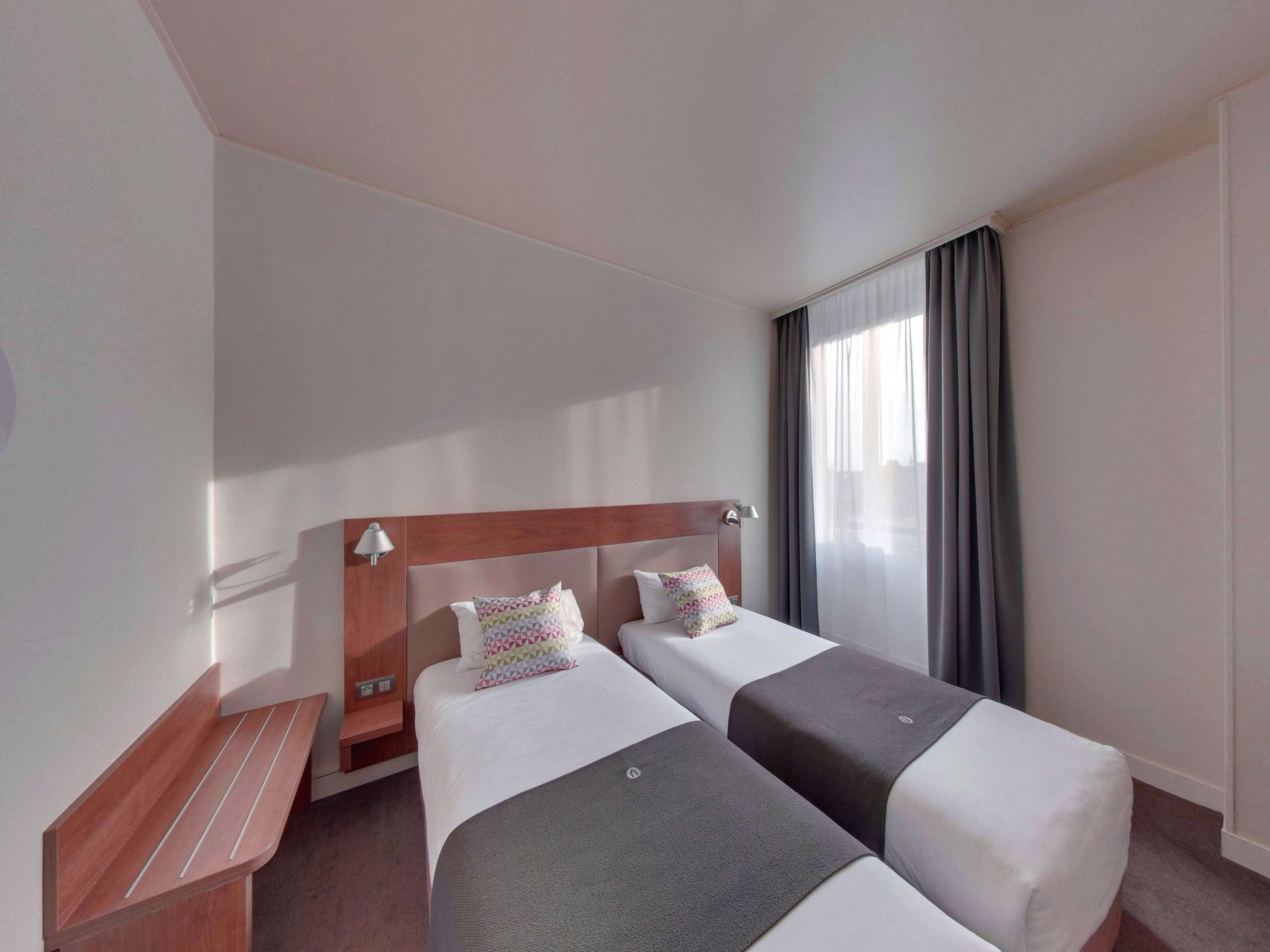 Habitación de hotel con dos camas, cabecero de madera, un banco y una ventana con cortinas.