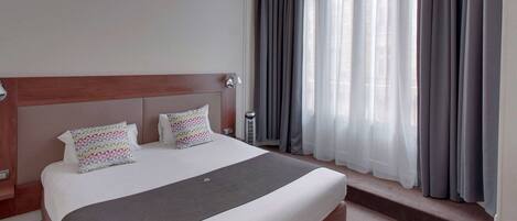 A hotel room with a large bed, a wooden headboard, a bedside table, a lamp, and a window with curtains.