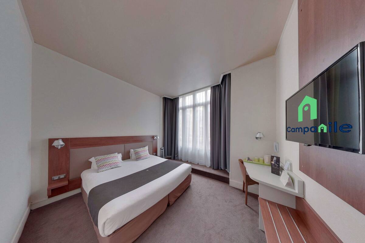 Una habitación de hotel con cama, escritorio, televisión y ventana con cortinas.