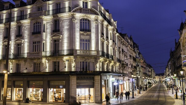 Un edificio histórico de varios pisos con detalles arquitectónicos ornamentados y un tranvía circulando por la calle.
