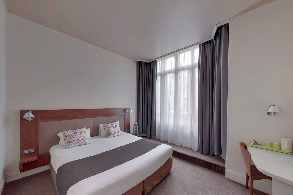 Habitación de hotel con una cama grande, una mesita de noche con lámpara, una ventana con cortinas y un escritorio pequeño con una silla.