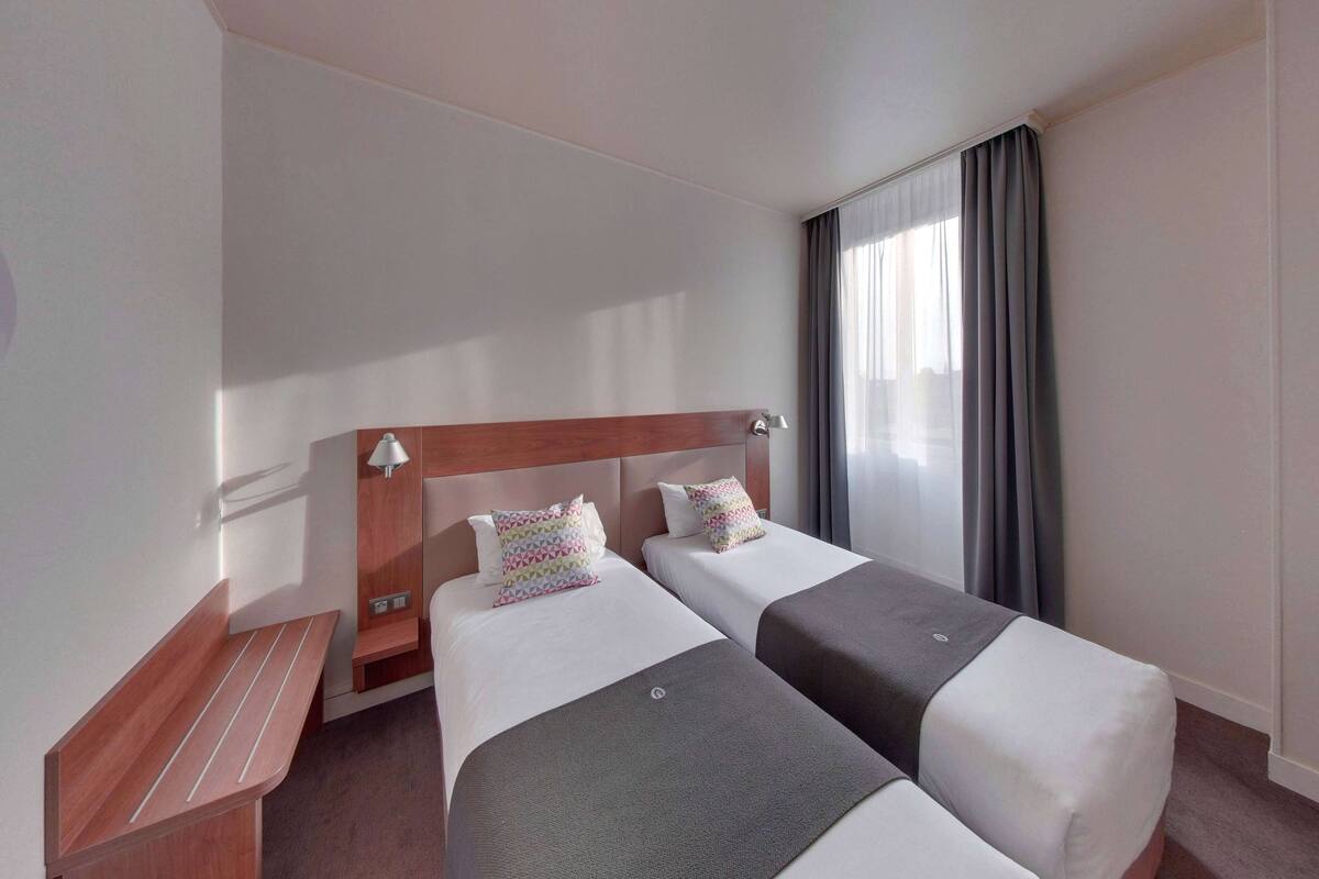 Habitación de hotel con dos camas individuales, cabecera de madera, mesita de noche y ventana con cortinas.