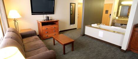 A hotel room with a flat-screen TV, a wooden dresser, a sofa, a small table, and a bathroom with a sink and mirror.