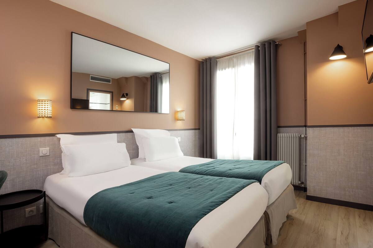 A hotel room with two beds, a large mirror, wall sconces, a window with curtains, and a radiator.