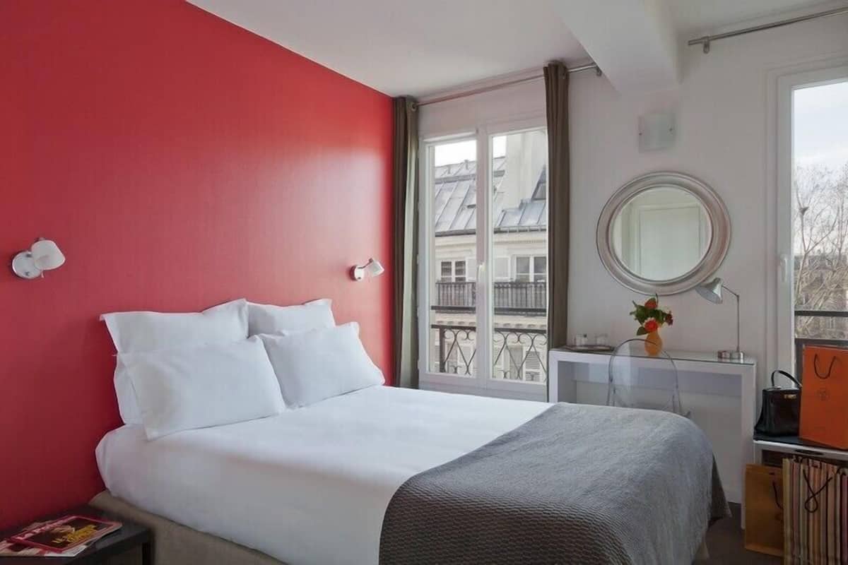 A hotel room with a bed, a bedside table, a mirror, and a window with a view of buildings.