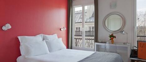 A hotel room with a bed, a bedside table, a mirror, and a window with a view of buildings.
