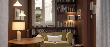 Un salón acogedor con un sofá, una mesita redonda, una estantería llena de libros y una ventana con vistas al verdor.