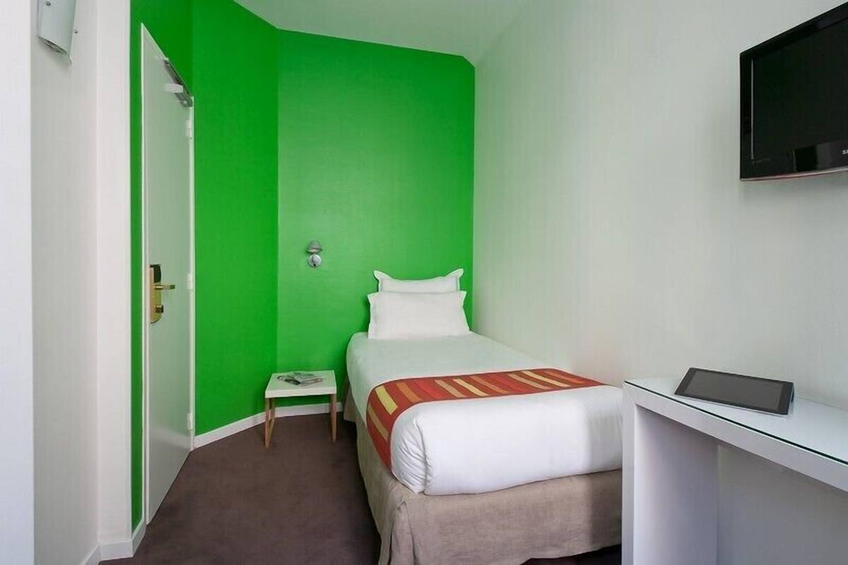 A hotel room with a bed, a small bedside table, a TV, and a green accent wall.