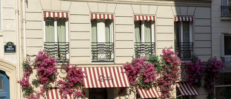 A hotel facade with a sign reading "HOTEL MARAIS DE LAUNAY," featuring multiple windows with red and white striped awnings, a blue door, and bicycles parked in front.