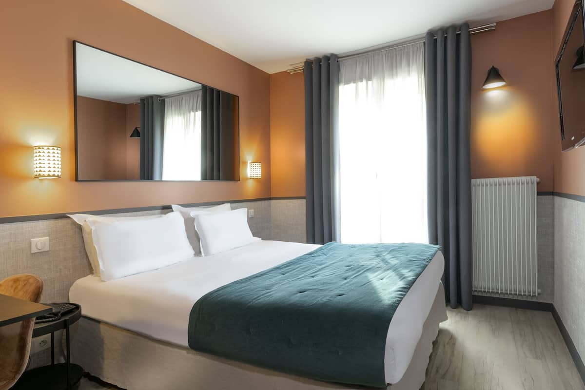 A hotel room with a bed, a desk, a chair, a mirror, and a window with curtains.
