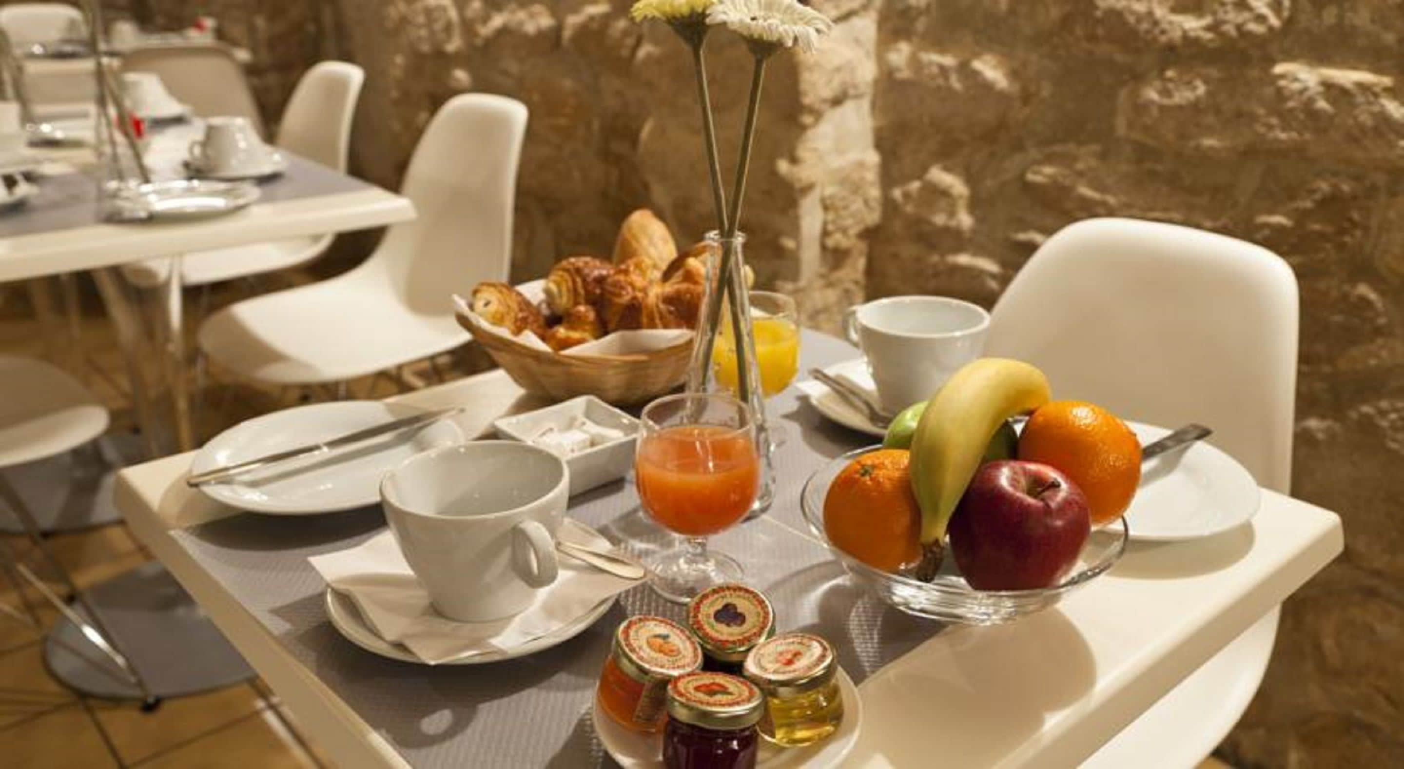 Une table mise pour le petit-déjeuner, avec une place pour deux personnes, comprenant des croissants, des fruits, du jus de fruits et de la confiture.