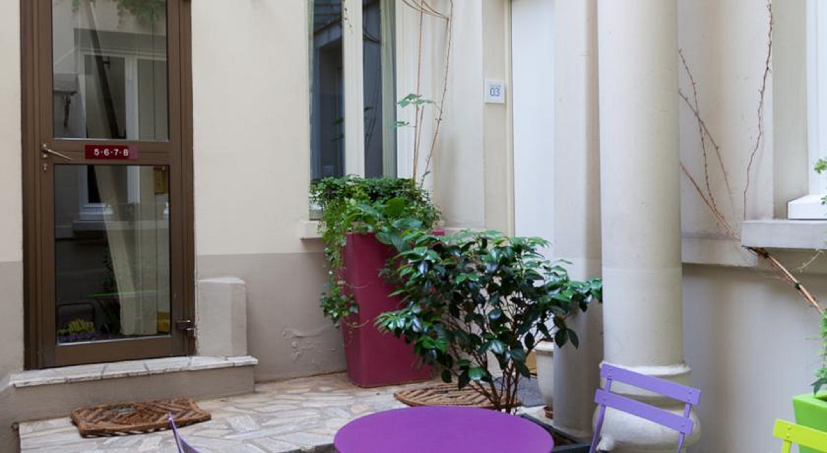 A small outdoor seating area with a purple table and a chair, a potted plant, and a door with a number sign.