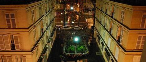Une vue nocturne d’une rue étroite bordée d’immeubles à étages et de balcons, qui mène à une place centrale où se trouvent un lampadaire et un petit arbre.