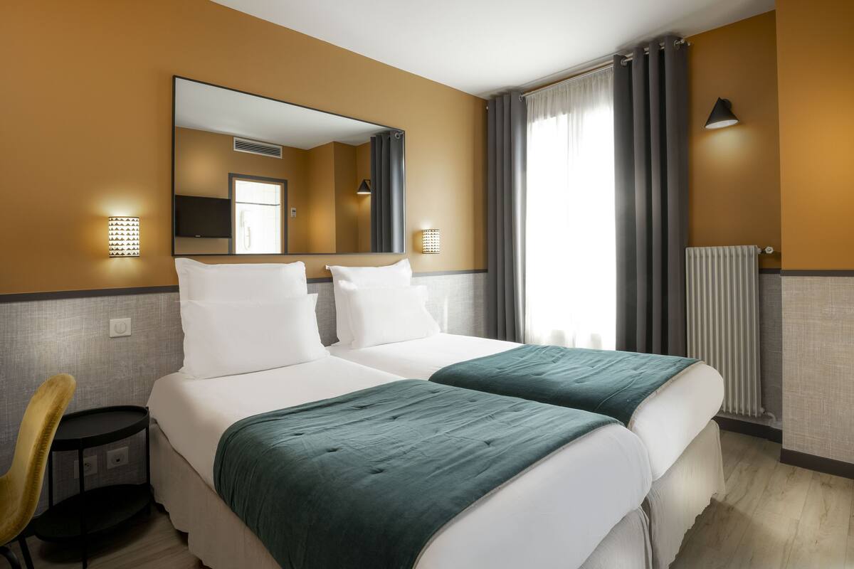 A hotel room with two beds, a chair, a mirror, and a window with curtains.