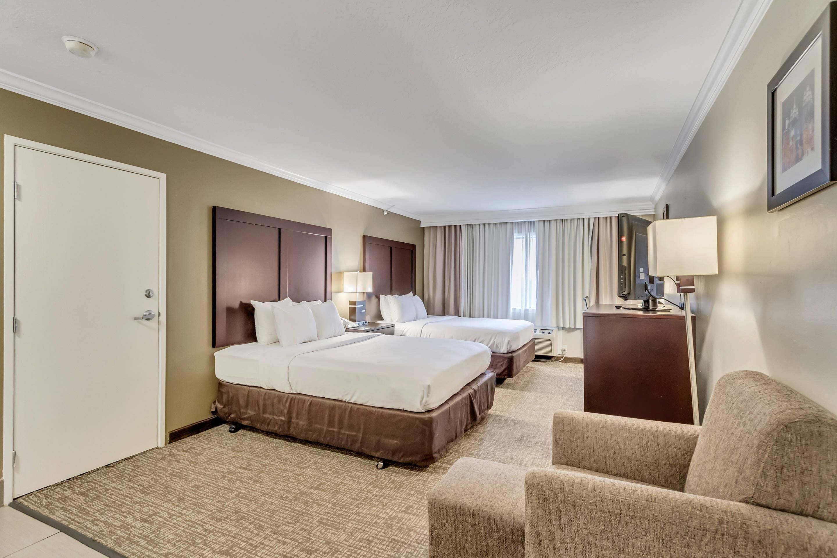 Habitación de hotel con dos camas, un escritorio, una silla y un ventanal grande con cortinas.