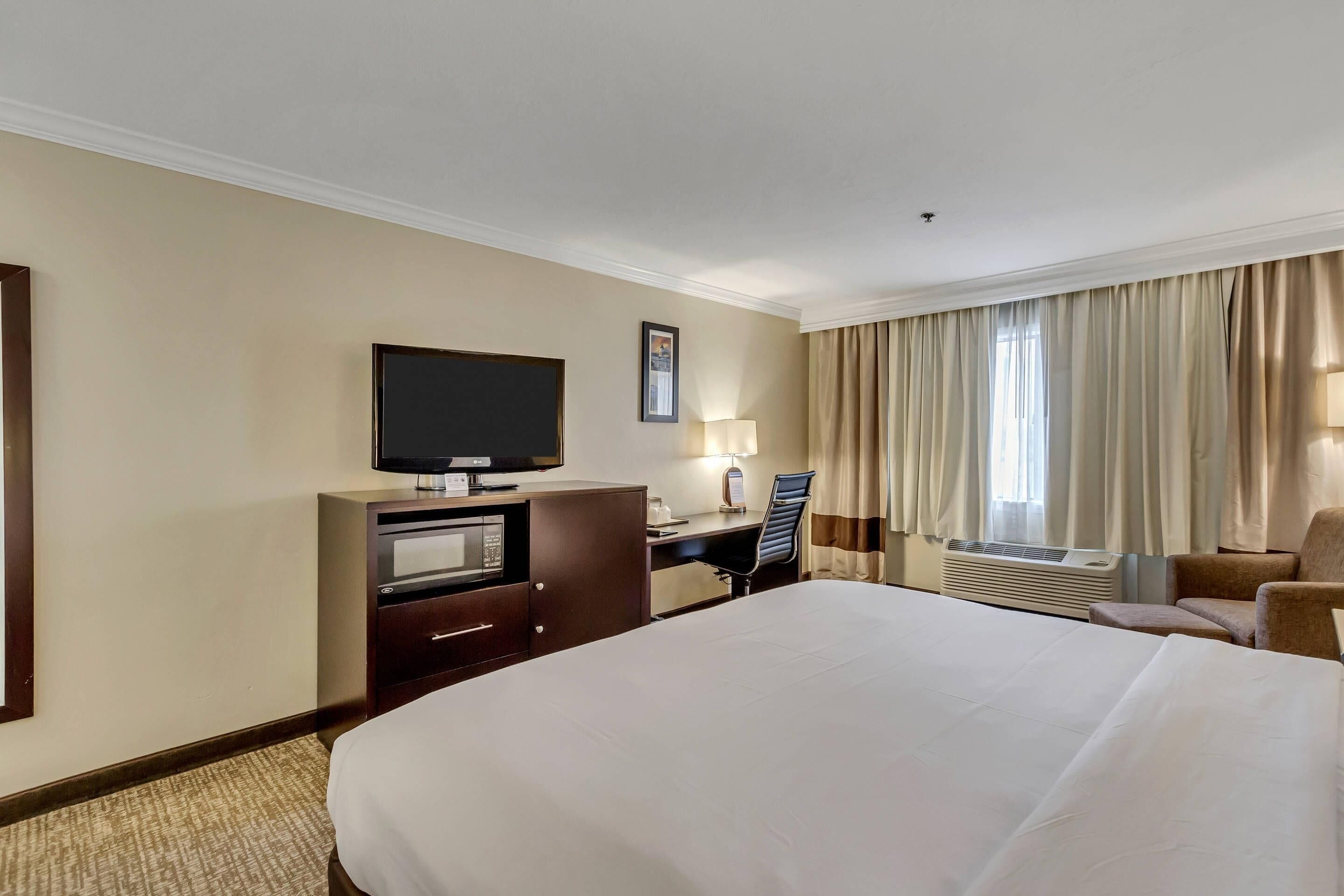 Une chambre d’hôtel avec un grand lit, un bureau avec une chaise, une télévision, un four à micro-ondes et une fenêtre avec des rideaux.