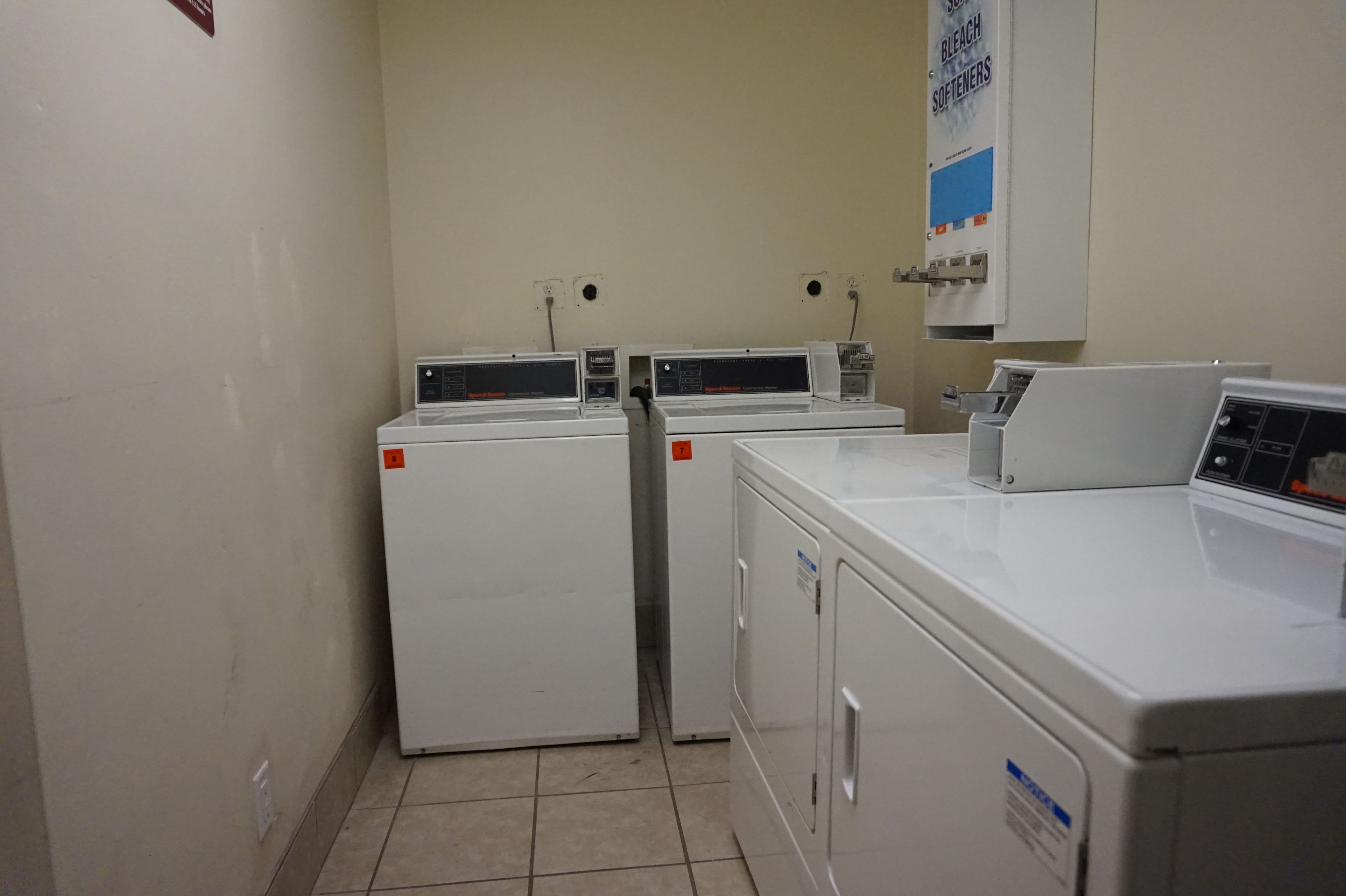 A laundry room with three stacked washers and dryers, a sign for bleach and softeners, and electrical outlets on the wall.