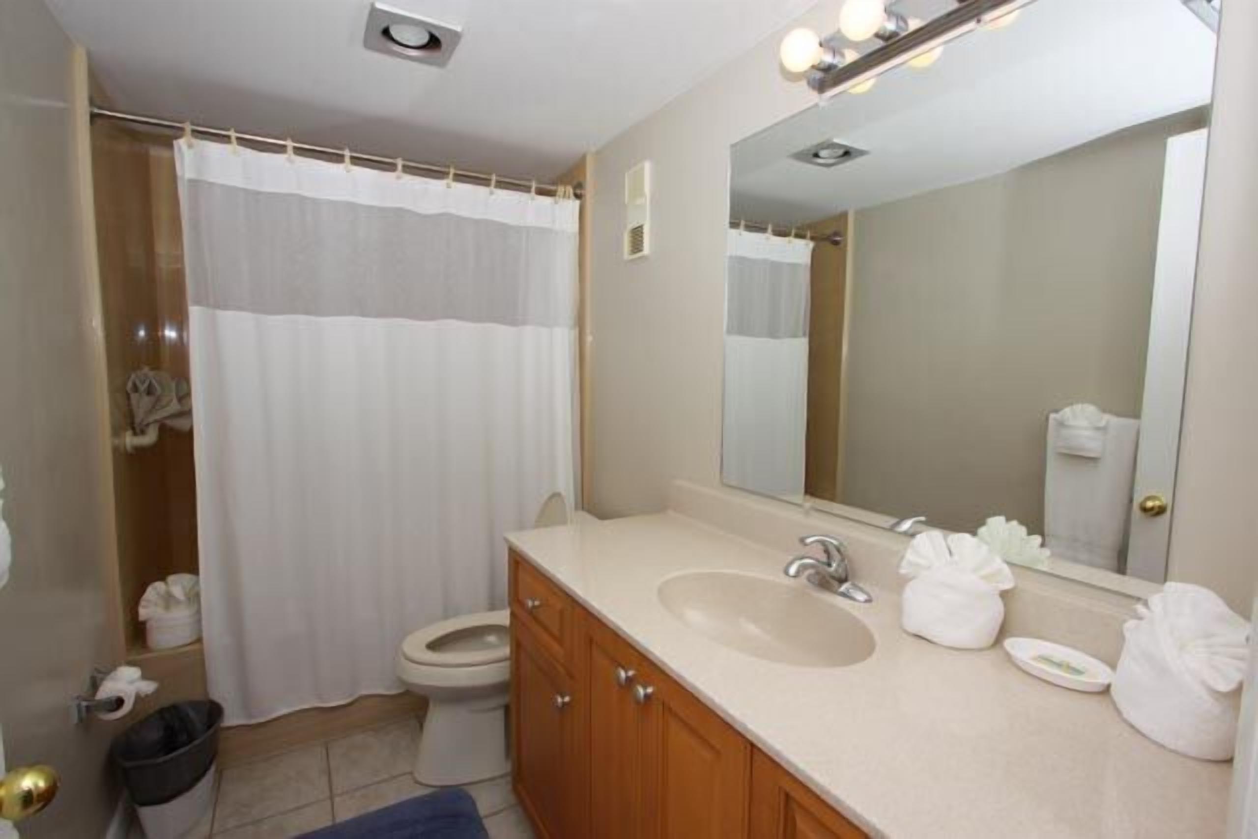 A bathroom with a shower curtain, toilet, sink, mirror, and a trash bin.