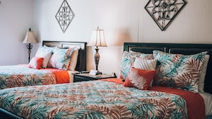 A hotel room with two beds, each with a floral-patterned bedspread and matching pillows. There are two bedside tables with lamps, and decorative wall art above the beds. - Cottages on the Green (Pensacola)
