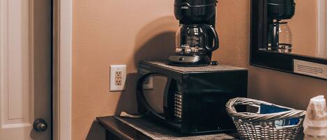 A kitchenette with a coffee maker, microwave, basket, and stacked cups.