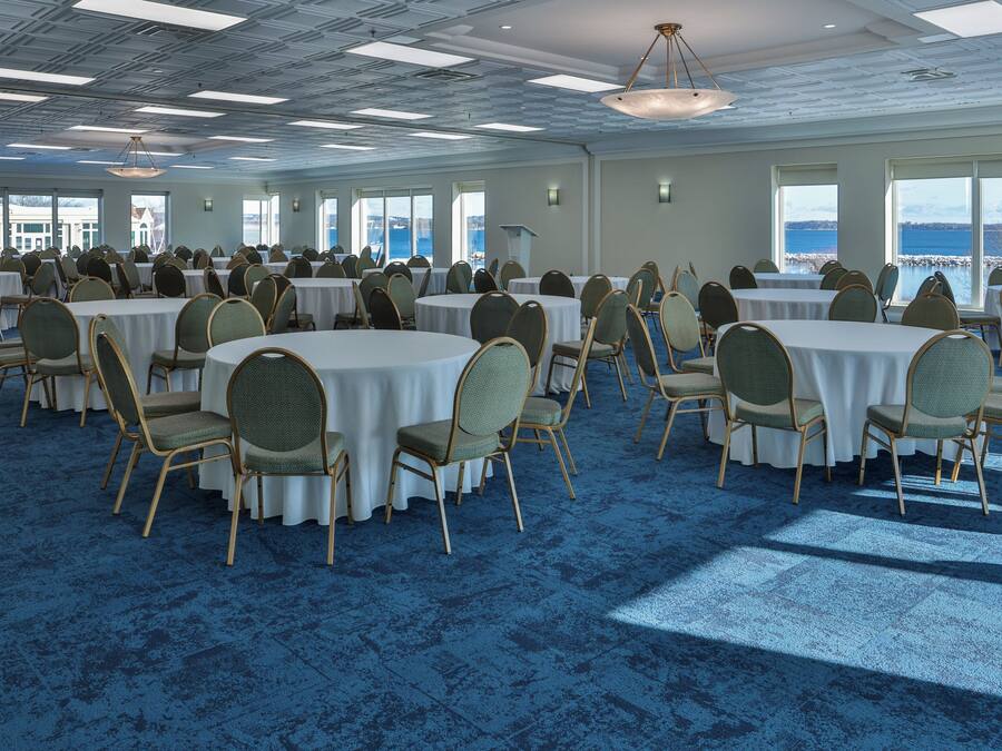 Une grande salle de conférence dotée de tables rondes et de chaises, de grandes fenêtres offrant une vue sur l’eau et d’un sol recouvert d’une moquette bleue.