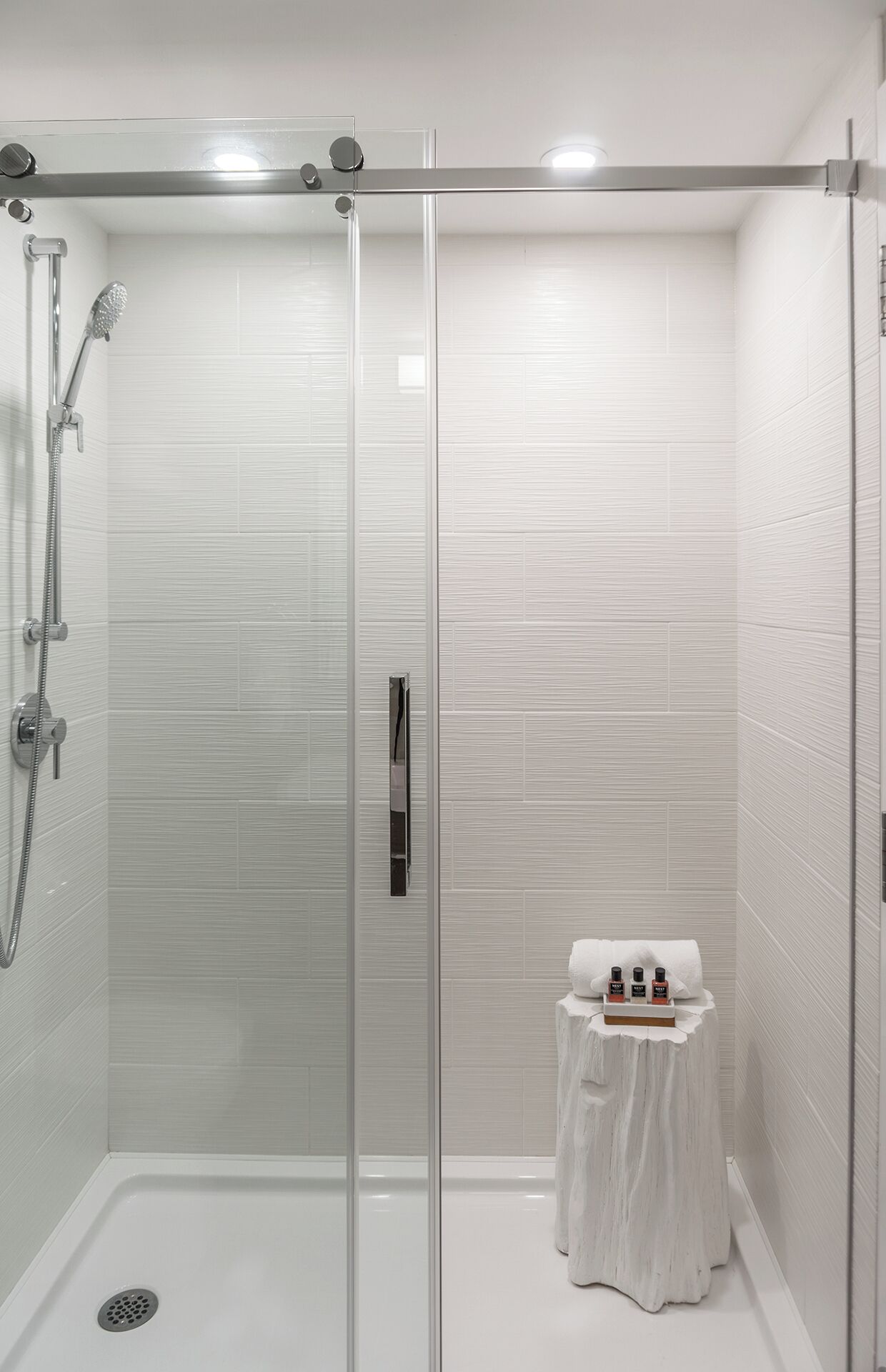A modern shower room with a glass door, a handheld showerhead, and a small table with toiletries.