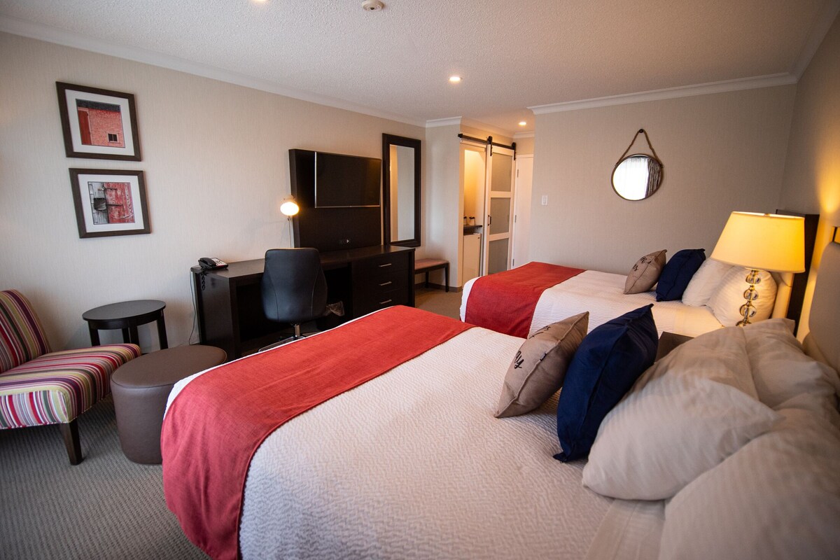A hotel room with two beds, a desk, a chair, a small stool, and a mirror.