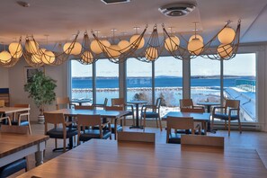 A modern dining area with wooden tables and chairs, large windows offering a view of a lake and bridge, and unique spherical hanging lights. - Oak Island Resort & Conference Centre (Western Shore)