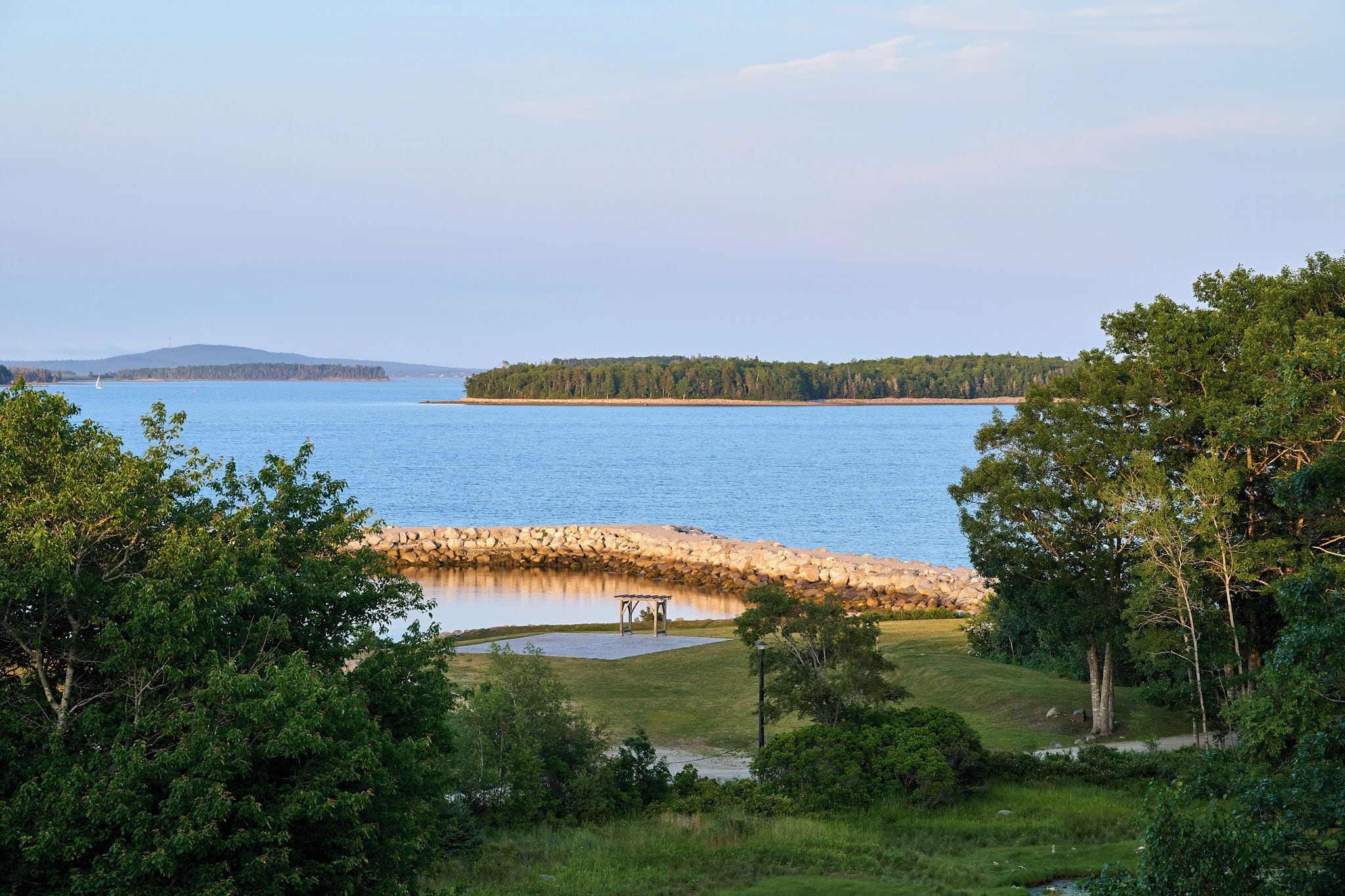 Blick auf einen See mit steiniger Uferlinie, einem Gazebo und dichter Vegetation.