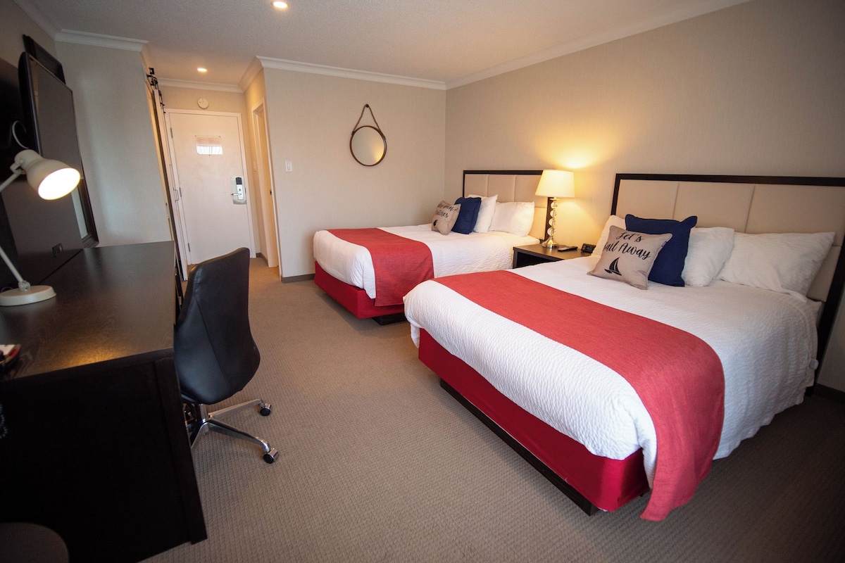 A hotel room with two beds, a desk with a chair, a desk lamp, and a wall-mounted mirror.