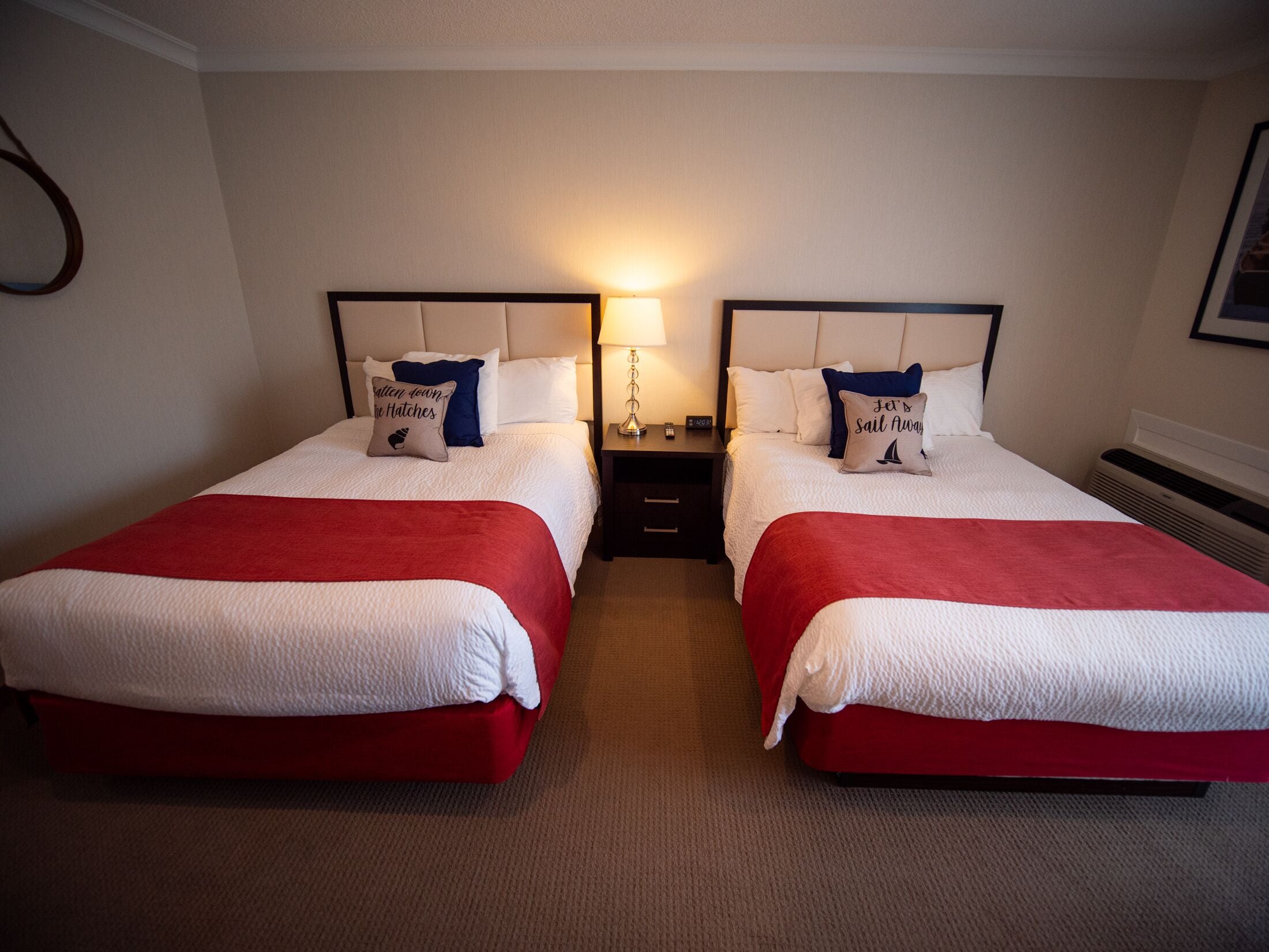 Camera d'albergo con due letti, ognuno con una coperta rossa e cuscini bianchi. Tra i letti, un comodino con una lampada.