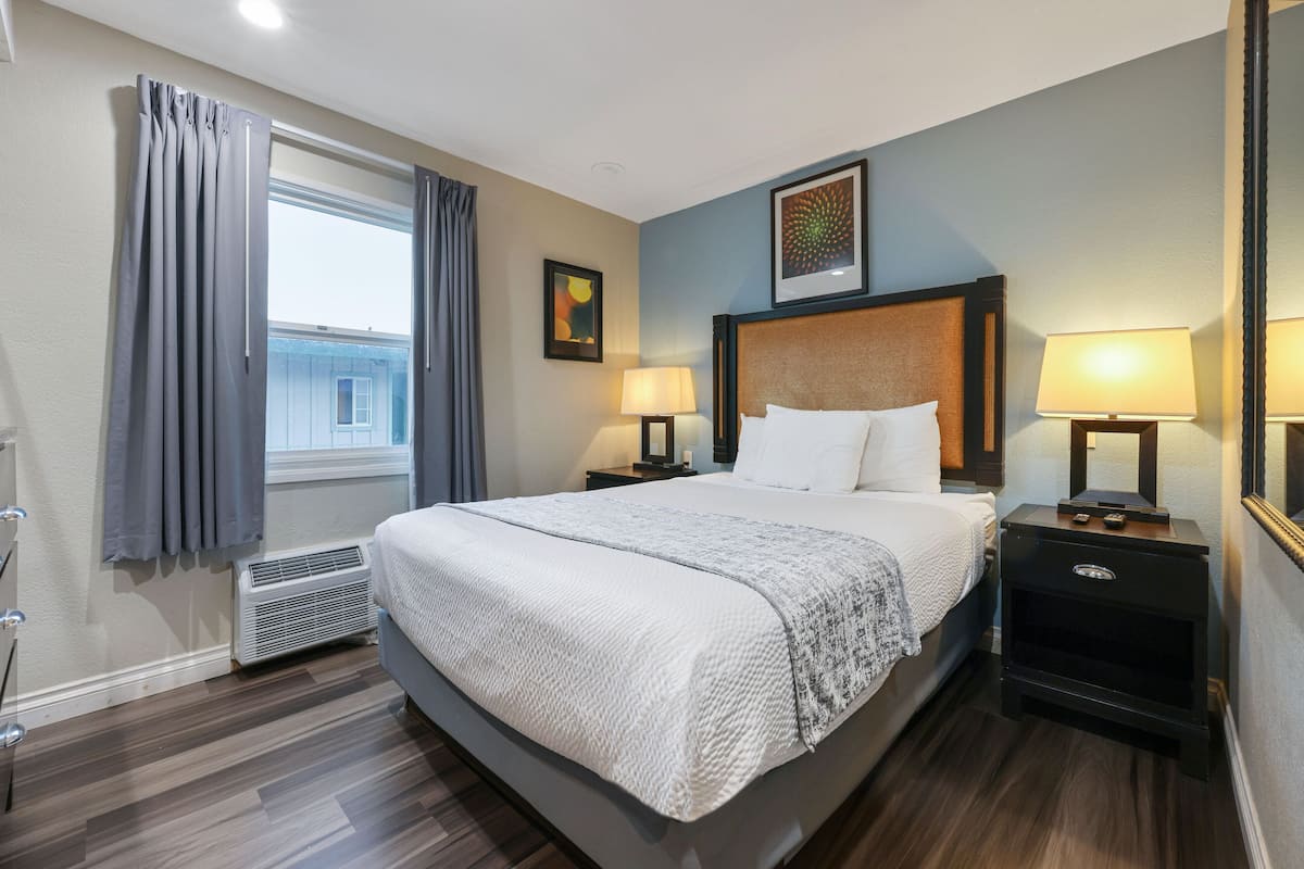 Habitación de hotel con una cama grande, dos mesitas de noche con lámparas, una ventana con cortinas, un radiador y un cuadro enmarcado en la pared.