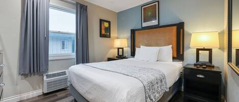 A hotel room with a large bed, two bedside tables with lamps, a window with curtains, a radiator, and a framed picture on the wall.