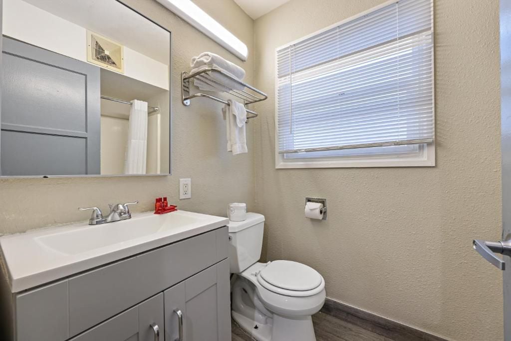 A bathroom with a white sink, mirror, and toilet. There is a towel rack above the toilet and a window with blinds.