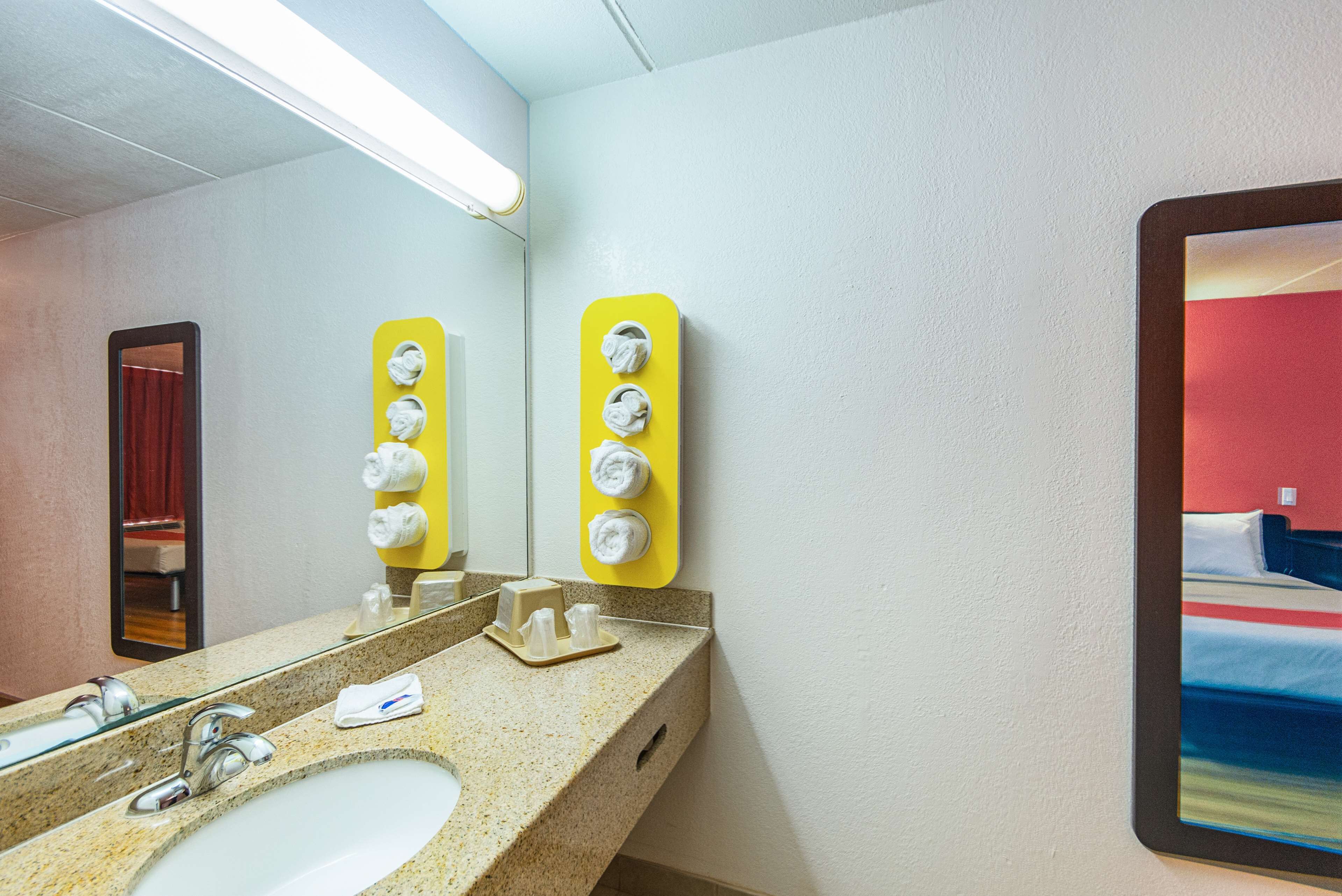 A hotel bathroom with a sink, mirror, towel dispensers, and a bed with a red wall in the reflection.