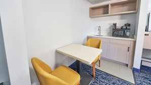 Cocina compacta con microondas, cafetera y una pequeña encimera. Hay una mesa con dos sillas amarillas y una sola silla. La cocina tiene armarios de madera clara y una alfombra azul estampada.