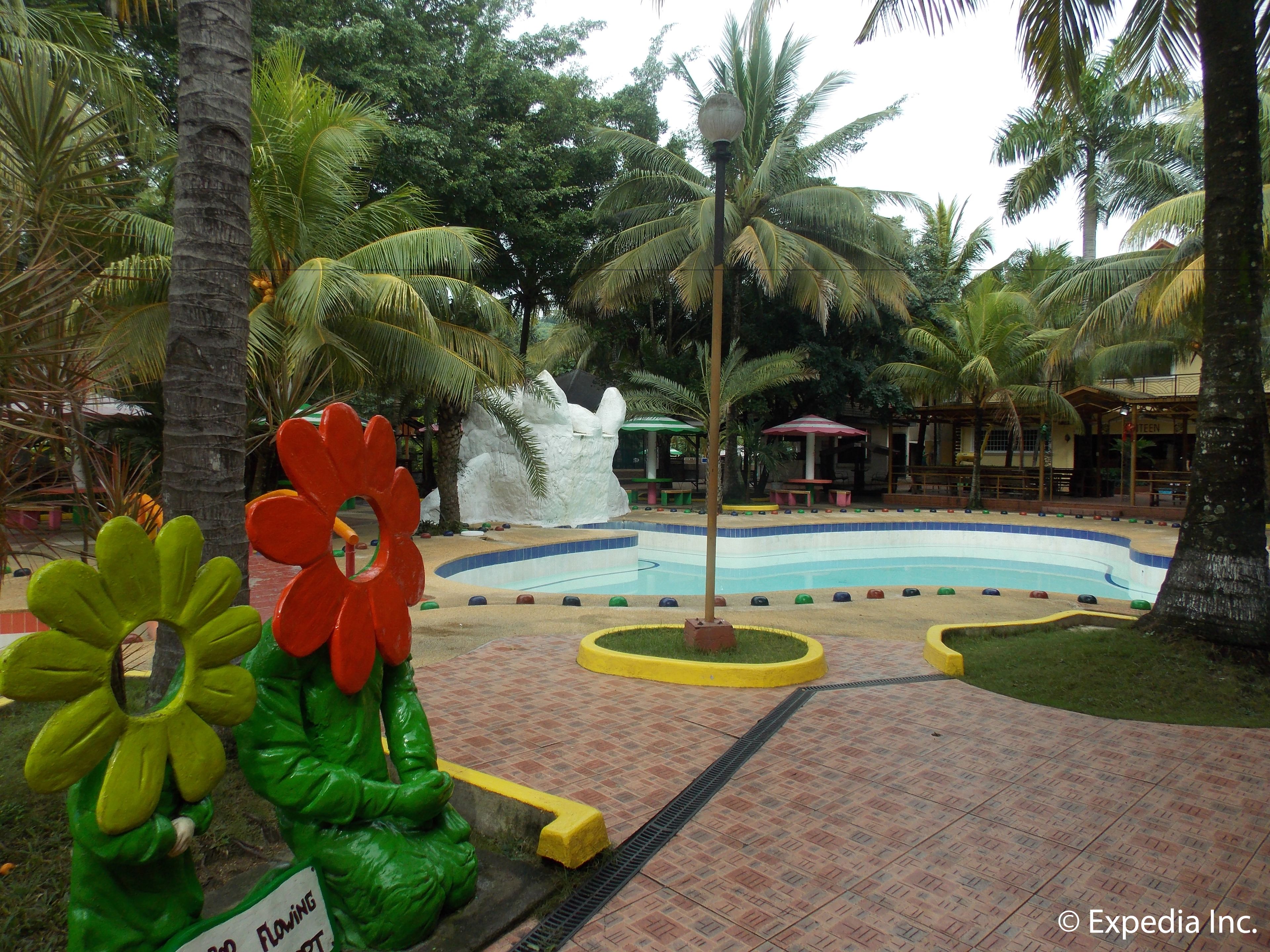 Un área de piscina con una fuente, palmeras y un parque infantil con estructuras con forma de flor.
