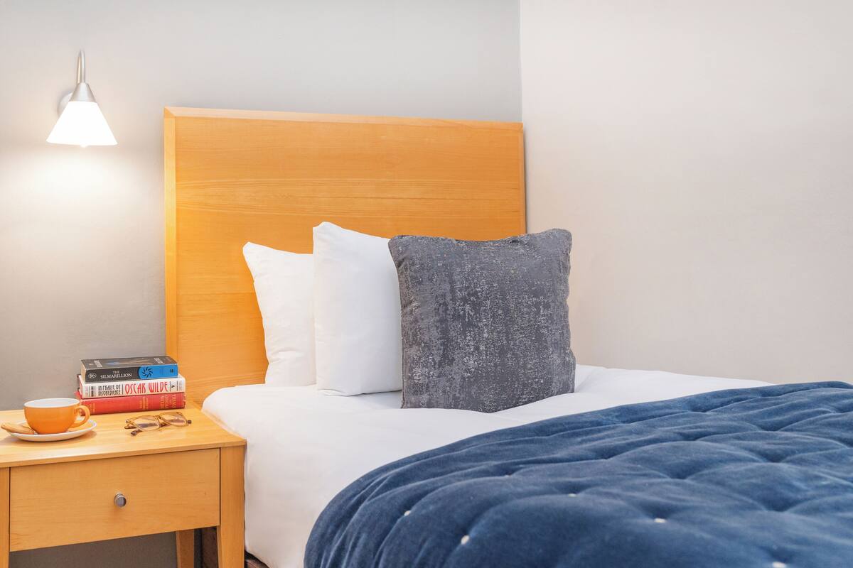 A neatly made bed with a wooden headboard, a blue blanket, and two pillows. There is a bedside table with a cup, a pair of glasses, and a stack of books. A wall-mounted lamp provides light.