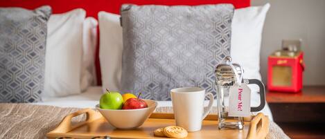 Bandeja de desayuno con un bol de fruta, una taza, una tetera y un periódico sobre una cama con almohadas estampadas y una pared roja.