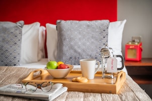 Bandeja de desayuno con un bol de fruta, una taza, una tetera y un periódico sobre una cama con almohadas estampadas y una pared roja.