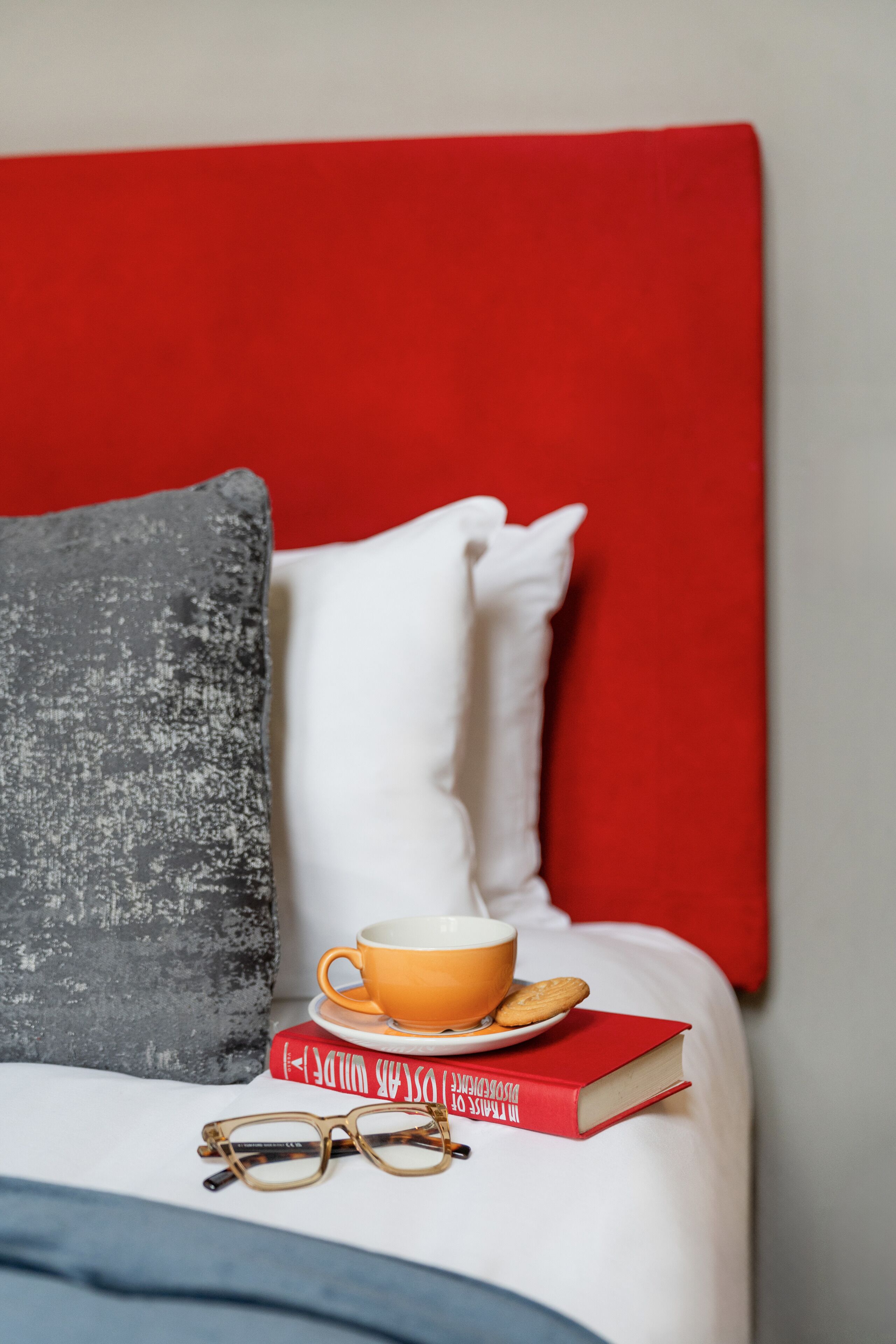 A bed with a red headboard, white pillows, a grey pillow, a cup, a book, and glasses.