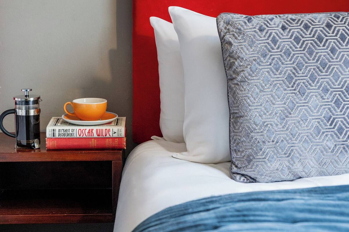 A neatly made bed with a red headboard, white pillows, and a patterned pillow. A bedside table holds a coffee maker, a mug, and a stack of books.