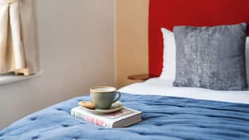 Una cama bien hecha con un cabecero rojo, una almohada gris y una manta azul. Hay una taza de café y un libro sobre la cama.