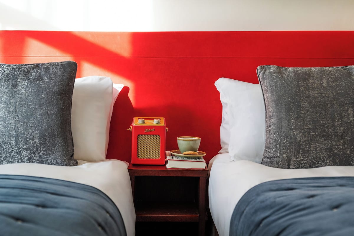 Two beds with grey pillows, a red radio, a cup, and a book on a nightstand.