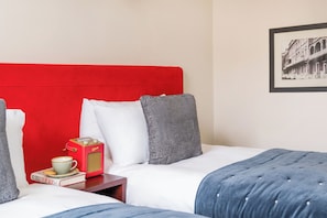 Habitación de hotel con cabecera roja, dos camas con ropa de cama gris y blanca, una mesita de noche pequeña con un libro y una taza, y un cuadro en la pared.