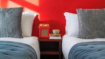 A room with two beds, each with a pillow and a blanket, a small table with a radio, a cup, and a book.