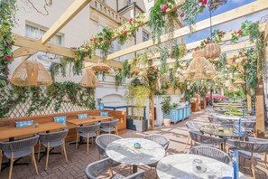 Un espacio al aire libre con mesas y sillas de madera, rodeado de abundante follaje y plantas colgantes.